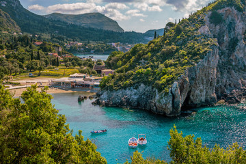 Beautiful landscape with sea–lagoon, beach, colorful boats on turquoise water surface, mountains and cliffs, green trees and bushes, blue sky and clouds. Corfu Island, Greece.  - obrazy, fototapety, plakaty