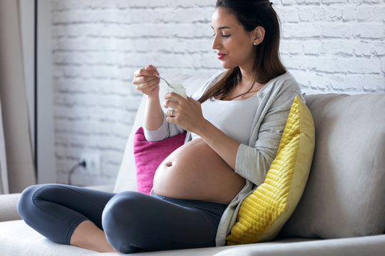 Beautiful pregnant young woman eating yogurt while sitting on sofa at home.