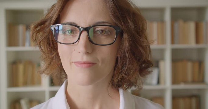 Closeup portrait of adult redhead attractive caucasian businesswoman looking at camera smiling cheerfully indoors with bookshelves on the background