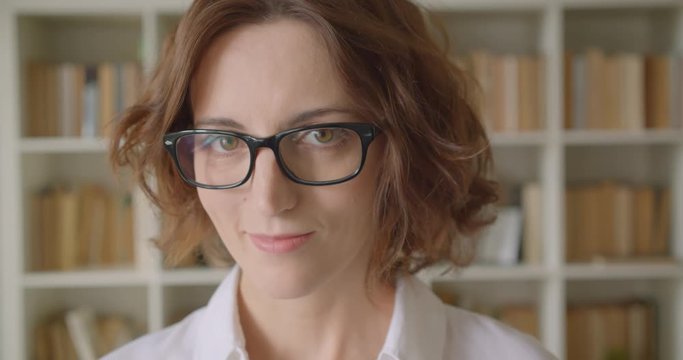 Closeup portrait of adult redhead beautiful caucasian businesswoman looking at camera smiling cheerfully indoors with bookshelves on the background