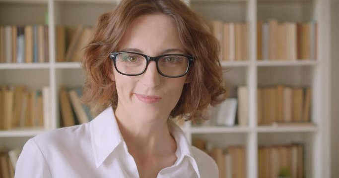 Closeup portrait of adult redhead beautiful caucasian businesswoman in glasses looking at camera smiling cheerfully indoors with bookshelves on the background