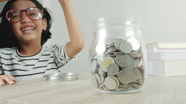Little Asian girl puttinwg the coin into piggy bank and smile with happiness for money saving to wealthness in the future of education concept select focus shallow depth of field