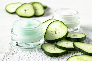 Face cream in a glass jar next to slices of fresh cucumbers on a white wooden background.