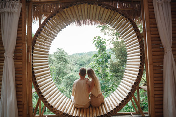 Back view couple sitting on the window in bamboo bungalow and looking at mountains. Vacation on Bali tropical island