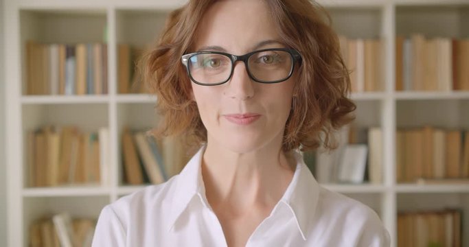 Closeup portrait of adult redhead caucasian businesswoman in glasses looking at camera smiling cheerfully indoors with bookshelves on the background