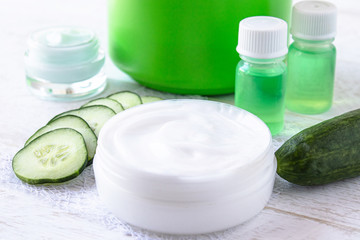 Face cream in a white jar with cucumber extract next to slices of fresh cucumbers on a white wooden background.