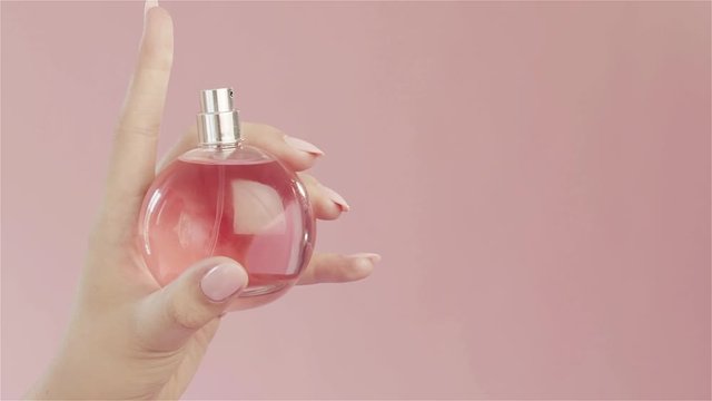 Beautiful tender graceful hand of girl with pink nails and long fingers, spray from  bottle of perfume with pink liquid in studio on pink background. Concept for advertising perfume.