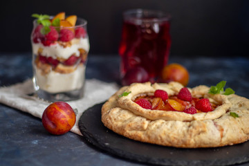 Biscuit with plums. Baked in the oven, next to a glass of cottage cheese cream with raspberries, fresh plums, all on a dark background. Near gray fabric and metal devices. 