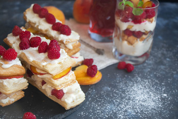 Dessert Milfey. Strips of puff pastry with curd cream raspberry berries and peach slices. In the background, the same, only in a glass. On a dark background. 