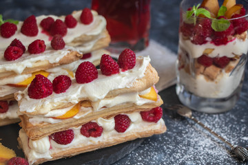 Dessert Milfey. Strips of puff pastry with curd cream raspberry berries and peach slices. In the background, the same, only in a glass. On a dark background. 