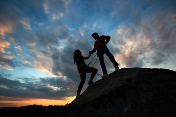 Two climbers reaching summit one holding hand of his partner assisting to make last step to top. Teamwork couple helping hand trust help, silhouette success in mountains. Amazing heaven on background.