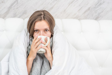 Beautiful sick woman sitting on the bed wrapped in a white blanket and drink hot tea. Empty space for text