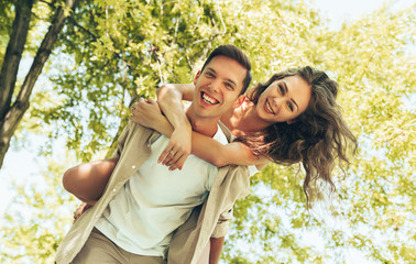 Portrait of young man carrying his beautiful woman on his back at the park, looking at camera....