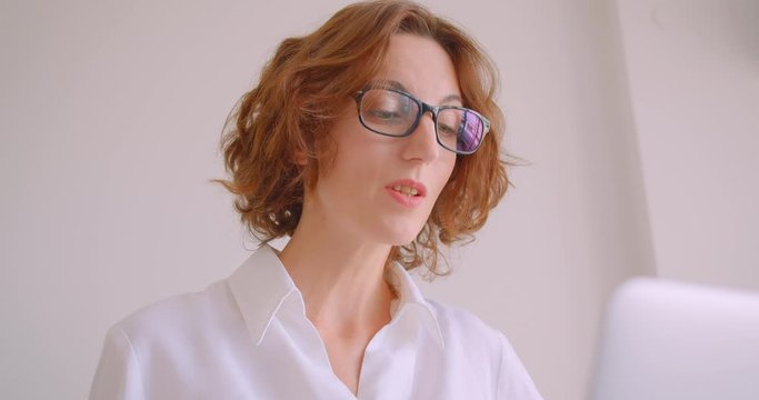 Closeup portrait of adult redhead caucasian businesswoman in glasses having a video call on the laptop sitting in the armchair in the white apartment