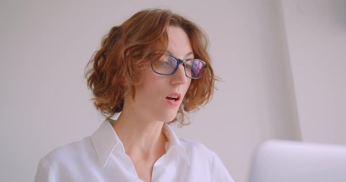 Closeup portrait of adult redhead caucasian businesswoman in glasses using the laptop being unhappy sitting in the armchair in the white office