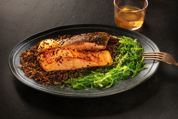 Grilled salmon with sesame seeds, wakame seaweed and wild rice, with a glass of white wine on a black background