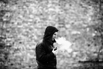 Vape man. Young caucasian white guy in black jacket with a hood on smoking and letting off steam...