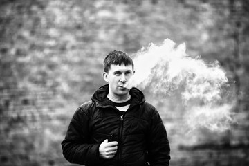 Vape man. Young caucasian white guy in black jacket smoking and letting off steam from an electronic cigarette opposite blurred background in the autumn. Bad habit. Vaping activity. Black and white