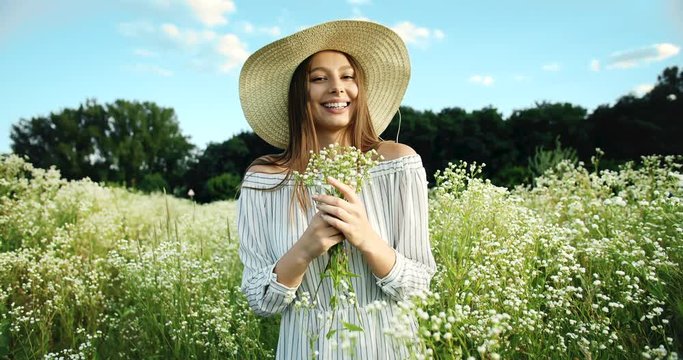 Attractive young lady in dress and hat smelling bunch of field flowers standing on green meadow, relaxing on nature