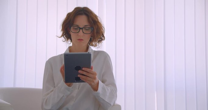 Closeup portrait of adult redhead caucasian businesswoman in glasses using the tablet looking at camera sitting in the armchair in the white office