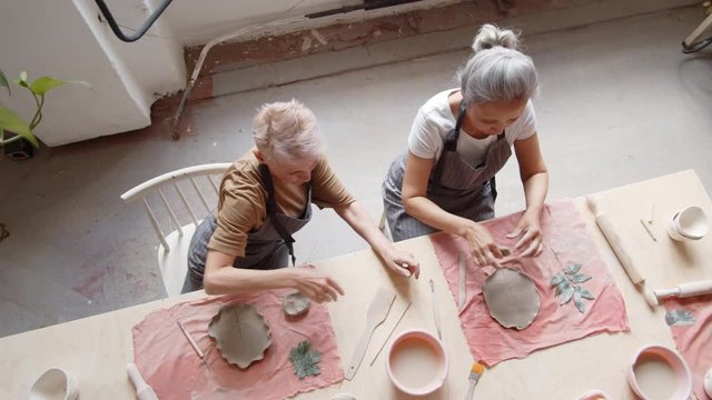 Top view of two Caucasian women sitting at wooden table in pottery workshop, communicating and decorating flat clay pieces using special tools