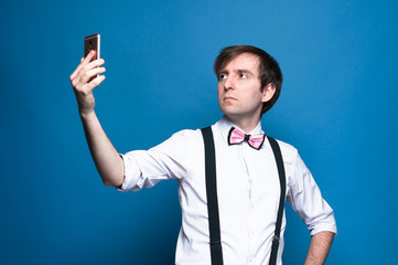 handsome man with dark hair in pink shirt with rolled up sleeves, bow tie and black suspender taking selfie on blue background with copy space