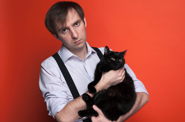 handsome bored man in shitr and black suspender holding cute black cat and looking at camera on orange background with copy space