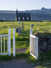 Black Church in Budir - PoI in western Iceland. The wooden church seen in late evening sun illumination through an open white painted wooden gate across an old cemetery. Mountains in background