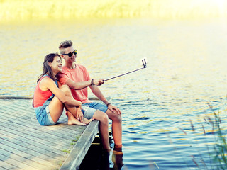 holidays, vacation, love and people concept - happy teenage couple taking picture by smartphone selfie stick and sitting on river berth at summer