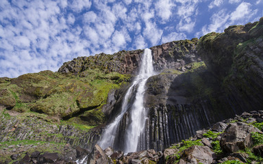 Summer in Iceland - view of a waterfall (Bjarnarfoss) in Snaefellsnes Peninsula. Stream falling from a high black volcanic rock formation. Above blue sky dotted by white clouds. Long exposure . 
