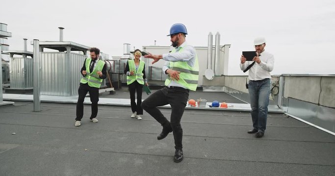 A bearded man dancing like a robot , and creating a fun atmosphere for the workers while they drink on a construction roof top