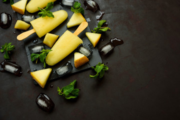 Homemade melon ice cream, pops or popsicles with ice, melon slices and mint on rectangular dark slate plate. Summer food. Flat lay. Top view