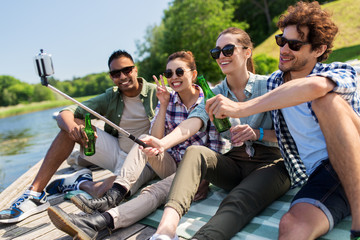 leisure, picnic and technology concept - friends with drinks taking picture by selfie stick on lake pier in summer park