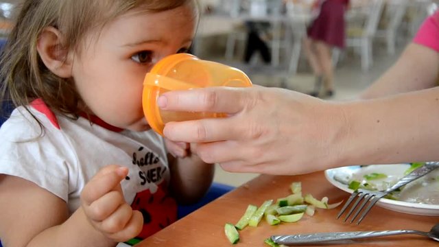 childhood and people concept - little baby drinking from spout cup sitting in highchair at home or at cafe dabbles, disobeys, builds faces, mom tries to feed