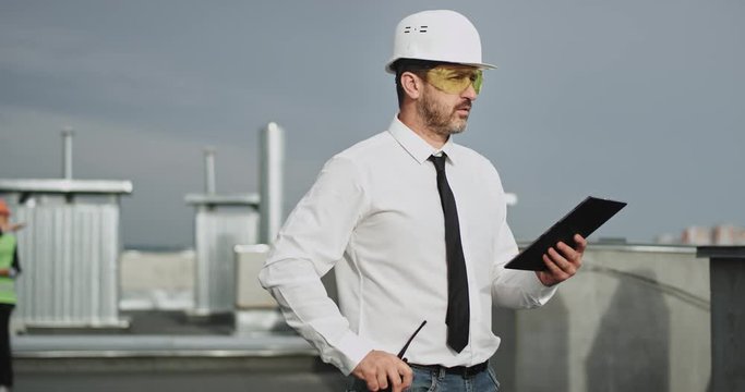 A handsome architect talks on his transceiver and puts it back and tapping on his iPad interested, while two people in the background are talking
