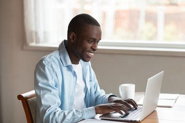African millennial guy sitting at table typing on laptop