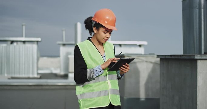 A tall beautiful young mixed woman , with Afro like hair tied back , talks on her transceiver while taking pictures with her iPad of the site , she looks very focused and ready to work