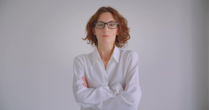 Closeup portrait of adult redhead caucasian businesswoman in glasses looking at camera with her arms crossed over chest indoors in the white apartment