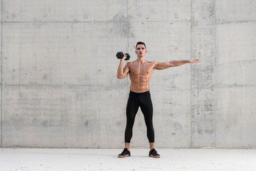 Muscular shirtless caucasian male athlete performs a dumbbell weightlifting exercise in a grungy concrete structure while showing his six pack abs 