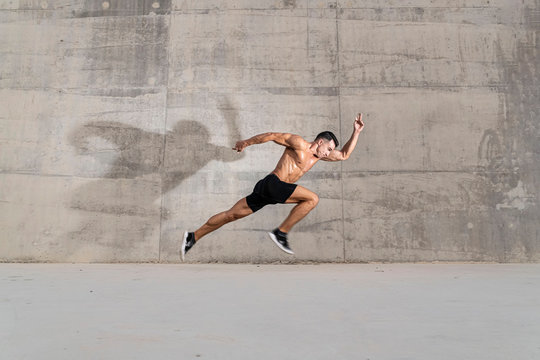 Muscular shirtless caucasian male athlete does an explosive starting sprint against  a grungy concrete wall background 