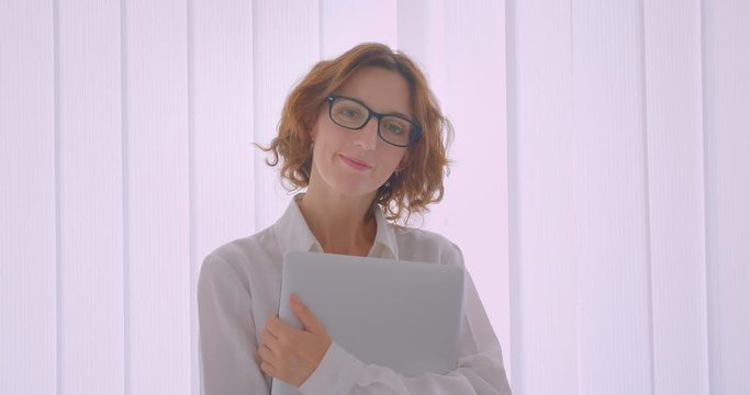 Closeup portrait of adult redhead caucasian businesswoman in glasses holding a laptop looking at camera indoors in the white apartment