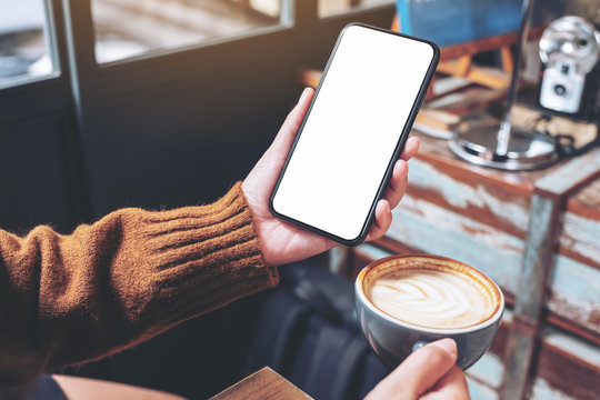 Mockup image of a woman holding black mobile phone with blank desktop screen while drinking coffee in cafe