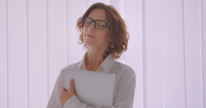 Closeup portrait of redhead caucasian businesswoman in glasses holding a laptop looking at camera smiling happily indoors in the white apartment