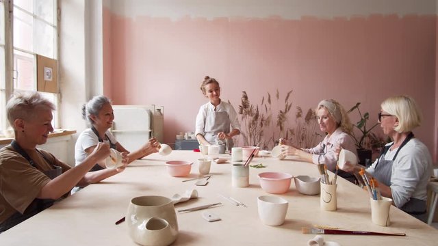 Zoom out of four middle-aged women in aprons sitting at wooden table in front of each other, painting clay cups with brushes while young tutor standing at table watching them