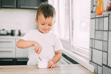 Cute boy in the kitchen eating yogurt from a white yogurt container, a place for advertising