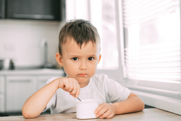 Cute boy in the kitchen eating yogurt from a white yogurt container, a place for advertising