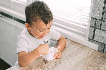 Fototapeta premium Cute boy in the kitchen eating yogurt from a white yogurt container, a place for advertising