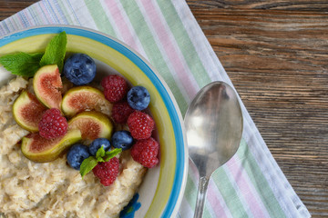 Delicious oatmeal, decorated with fig pieces, raspberries and blueberries, and mint leaves is in a bowl on a wooden table on a napkin next to a spoon, top view, close-up
