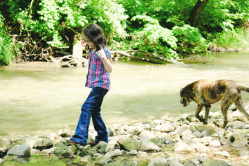 Child outdoors in nature shows young girl walking by creek in woods with pet dog.
