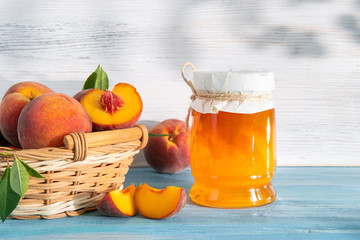 Jam and peaches with seeds and leaves in a wicker basket on a blue wooden background and on a white brick wall. The jar is covered with canvas.